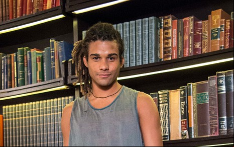 O ator Lucas Leto, com uma regata cinza e dreadlocks presos, posa em frente uma estante cheia de livros caracterizado como Waguinho em cena de Bom Sucesso
