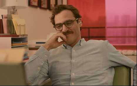 Joaquin Phoenix caracterizado como Theodore em Ela, de camisa azul clara e óculos marrom, o ator apoia o rosto sobre sua mão, com feição pensativa, ele esboça um tímido sorriso