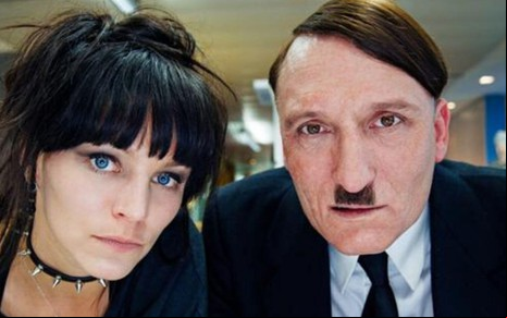 Em cena do filme Ele Está de Volta, os atores Franziska Wulf e Oliver Masucci olham para a câmera, curiosos