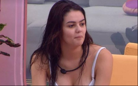Eslô veste blusa branca e está olhando para o lado; ela está sentada em um sofá amarelo