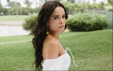 Caroline Macedo posa de perfil para foto no meio de um extenso gramado; ela tem o semblante sério enquanto encara a câmera, usando um vestido branco. Os cabelos longos e ondulados estão soltos