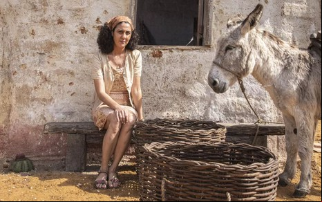 A atriz Clarissa Pinheiro como Tereza em Mar do Sertão; ela está sentada, com um lenço na cabeça, e as mãos nas pernas, ao lado de um burro e um balaio