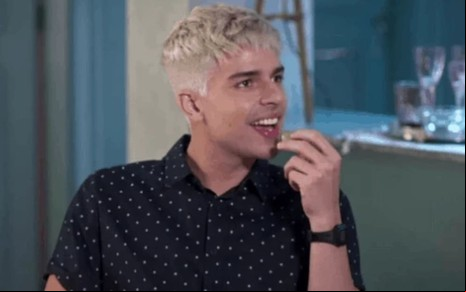 Diego Montez tem os cabelos platinados e veste uma camisa polo preta; ele sorri, ironicamente, em cena de Bom Sucesso