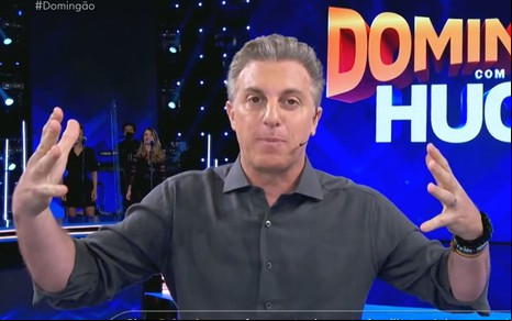 Luciano Huck abre os braços enquanto apresenta o Domingão nos Estúdios Globo; ele está de camisa cinza