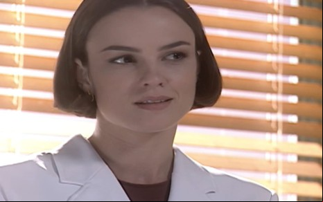 Carolina Kasting caracterizada como Laura; ela usa um jaleco branco e os cabelos curtos, na altura do queixo. A atriz dá um sorriso cúmplice em cena de Mulheres Apaixonadas