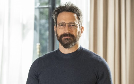 O ator Caco Ciocler, com barba, óculos e camiseta escura, posa no consultório do seu personagem na novela Pantanal