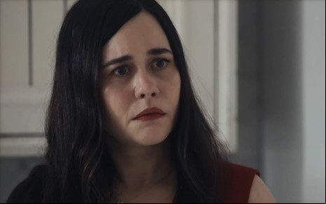 Alessandra Negrini caracterizada como Guida em Travessia; ela tem o semblante abalado e usa uma blusa vermelha em cena da novela