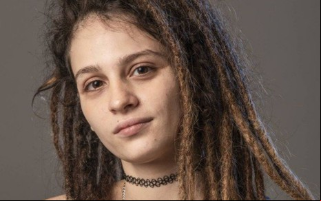 Lara Tremouroux está caracterizada como Joy em foto de divulgação de Um Lugar ao Sol; ela está em frente à parede cinza