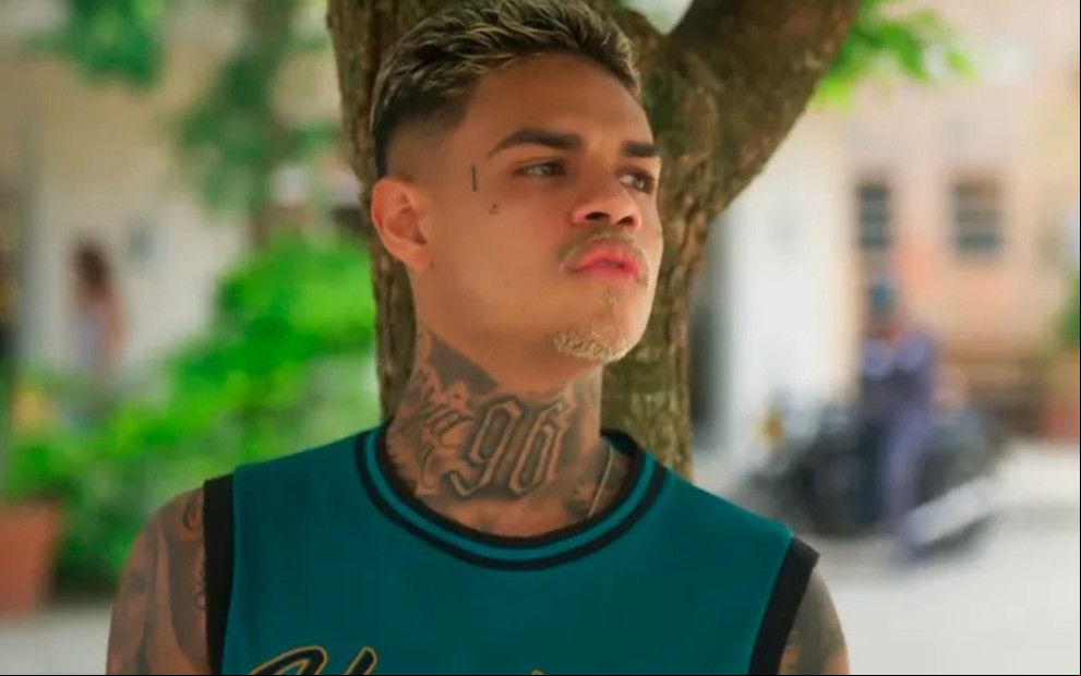 O ator MC Cabelinho com expressão séria, na frente de uma árvore em cena de Vai na Fé