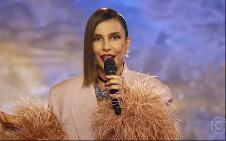 Ivete Sangalo com um vestido bege e sorrindo para a câmera no cenário do Masked Singer Brasil