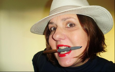 Jackie Vellego está de chapéu bege, ela segura uma caneta com os dentes e usa batom vermelho