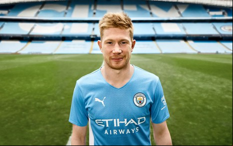 Kevin De Brunyne com a camisa azul clara do Manchester CIty, sorrindo e mostrando o estádio Etihad Stadium, em Manchester (ING)