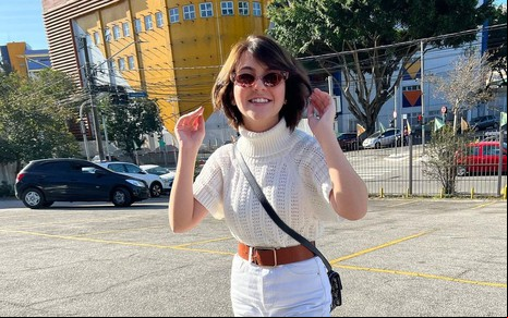 A atriz Klara Castanho; ela está usando um óculos de sol, sorrindo e com as mãos para cima na altura do ouvido