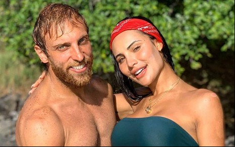 Leo sorri, está sem camisa e abraça Aline; Aline sorri, usa bandana vermelha e top verde