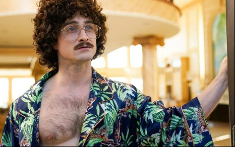 Com uma camisa florida aberta que mostra o peito peludo, Daniel Radcliffe usa um bigode falso e uma peruca encaracolada