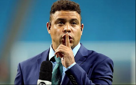 Ronaldo Fenômeno com um terno azul, gravata verde e um sinal de silêncio durante cobertura de um jogo na Seleção Brasileira