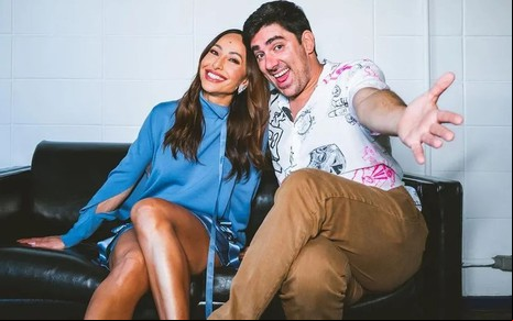 Sabrina Sato e Marcelo Adnet estão sentados em um sofá preto; no fundo há uma parede branca: ela está com vestido azul e ele com calça marrom e blusa estampada