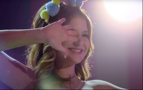 A atriz Sophia Valverde, caracterizada como a personagem Poliana, sorri com a mão esquerda na frente de seu rosto na novela Poliana Moça, do SBT