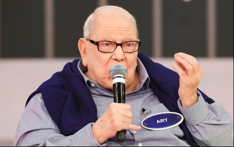 Ary Toledo usa uma blusa social preta e um casaco azul marinho; ele segura o microfone perto da boca