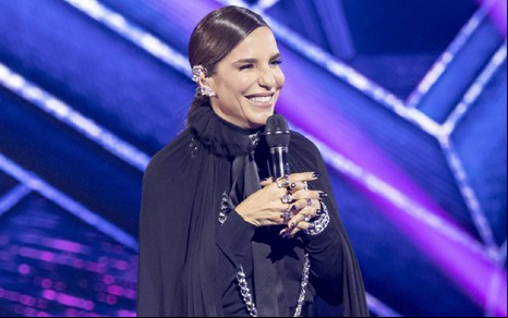 Ivete Sangalo com um vestido preto e sorrindo para a câmera no cenário do The Masked Singer Brasil. O fundo é azul.