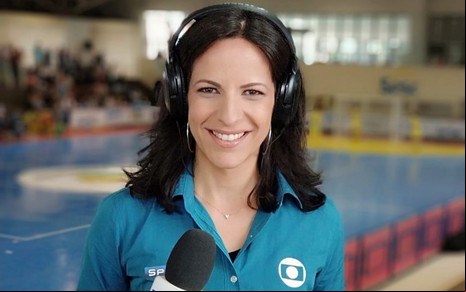 Viviane Costa com uma camisa azul, segurando o microfone da Globo em uma arena esportiva