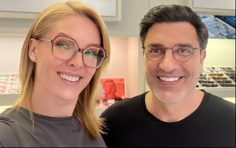 Ana Hickmann veste uma blusa cinza, e Edu Guedes, uma preta; os dois usam óculos e sorriem para a câmera