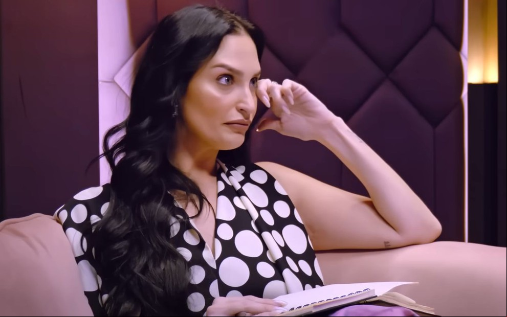 Ariela Carasso está sentada em sofá e parece enxugar uma lágrima discretamente