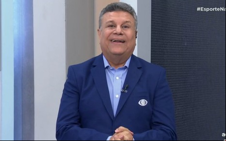 Téo José sorri em cenário esportivo da Band; ele usa paletó azul escuro e camisa social clara