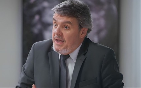 O ator Cassio Gabus Mendes com expressão assustada, de boca aberta, de terno e gravata, em cena de Elas por Elas