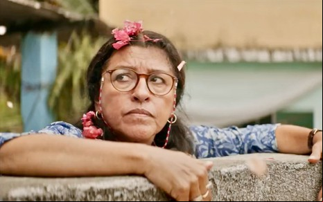 A atriz Regina Casé está de óculos apoiada em um muro em cena de Dona Lurdes, o Filme