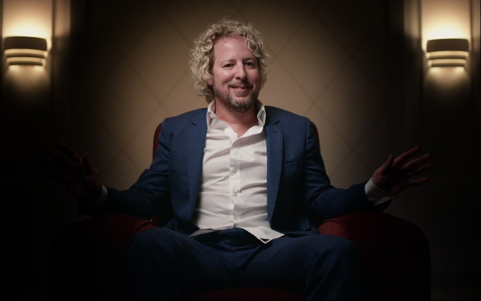 Jonny Fairplay está sentado em uma poltrona em uma sala pouco iluminada; os braços e as pernas dele estão na escuridão, enquanto ele força um sorriso