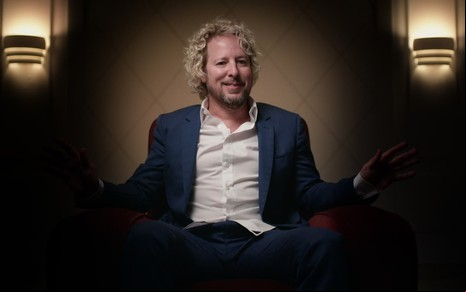 Jonny Fairplay está sentado em uma poltrona em uma sala pouco iluminada; os braços e as pernas dele estão na escuridão, enquanto ele força um sorriso