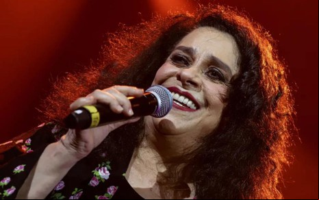 Gal Costa se apresenta no palco, com um microfone nas mãos, e sorri