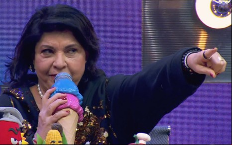 A cantora Roberta Miranda está com o dedo da mão direita apontado para a direita segurando um microfone em formato de sorvete no Caldeirão