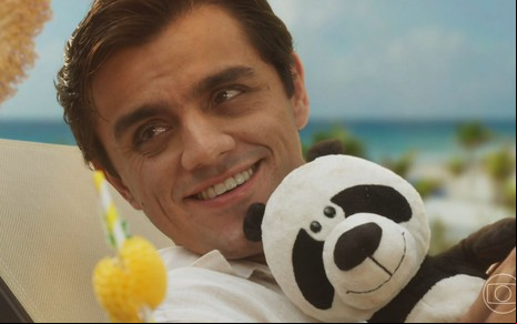 Um homem sorridente, deitado em uma espreguiçadeira, segura um ursinho de pelúcia estilo panda, com um cenário de praia ao fundo.