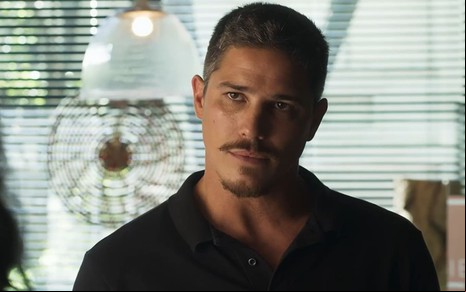 Romulo Arantes Neto caracterizado como Julião; ele usa uma camisa preta e está sério em cena de Fuzuê