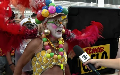 Um homem de cerca de 70 anos está vestido como uma drag queen, com maquiagem branca e bolas coloridas na Banda de Ipanema, no Rio