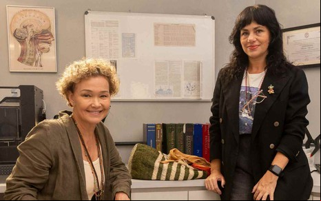 Julia Lemmertz e Karine Teles em Elas por Elas, Julia sentada e Karine de pé, ambas sorrindo e olhando para a câmera
