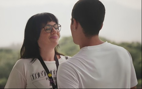 Carol (Karine Teles) olha sorridente para Marcos (Luan Argollo), que aparece de costas em cena da novela Elas por Elas