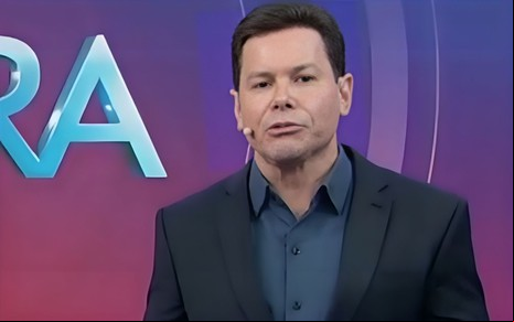 Um apresentador de cabelos escuros veste terno preto e camisa azul. Ele fala em frente a um fundo roxo com letras grandes e brilhantes.
