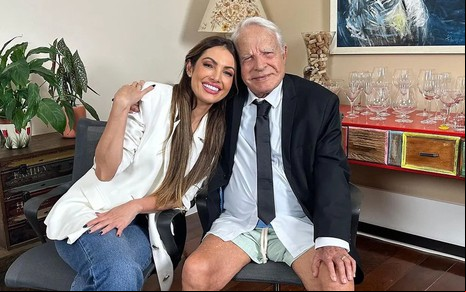 Patrícia Poeta de blazer branco sorri ao lado de Cid Moreira, que está de paletó, gravata e short