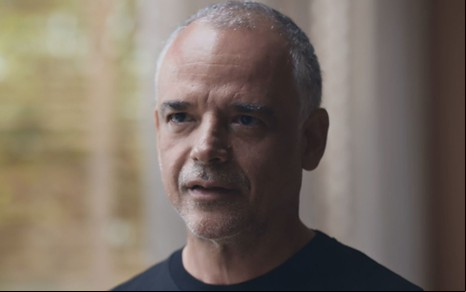 Ângelo Antônio usa uma camiseta preta e está com o cabelo bem curto, quase raspado; ele está sério em cena de Todas as Flores