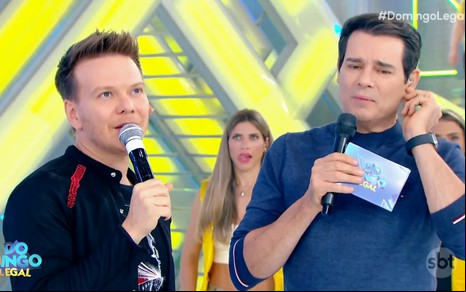 Michel Teló fala ao microfone enquanto Celso Portiolli faz careta no palco do Domingo Legal
