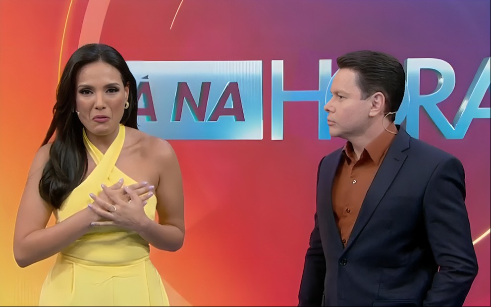 Márcia Dantas leva as mãos ao peito e aparenta emoção no cenário do Tá na Hora; Marcão da Povo a observa
