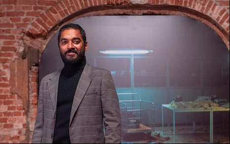 Thomás Aquino usa blusa preta de gola alta e um blazer cinza; no fundo há uma parede de tijolinho