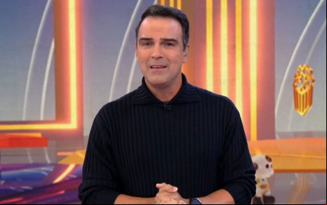 Homem de cabelo curto veste suéter preto em estúdio colorido, com detalhes amarelos e vermelhos ao fundo.