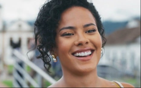 Mulher sorridente com cabelo cacheado preso, usando brinco dourado com pérola, em ambiente ao ar livre com construções ao fundo.