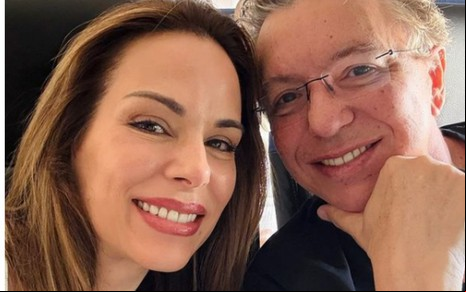 Duas pessoas sorrindo em um close. À esquerda, uma mulher de cabelos castanhos longos e maquiagem leve; à direita, um homem usando óculos.