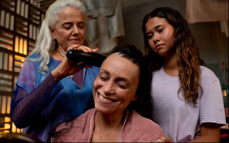 A atriz Marieta Severo raspando a cabeça da atriz Suzana Pires, que está sorrindo, e a atriz Nathalia Costa olhando, séria