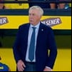 Técnico de terno azul e gravata azul claro em frente a placa amarela da Pilsener durante partida de futebol.